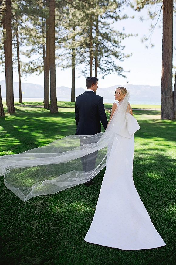 Couple portrait of bride and groom walking away holding hands, cathedral veil blowing by a mountain lake with pine trees and blue sky