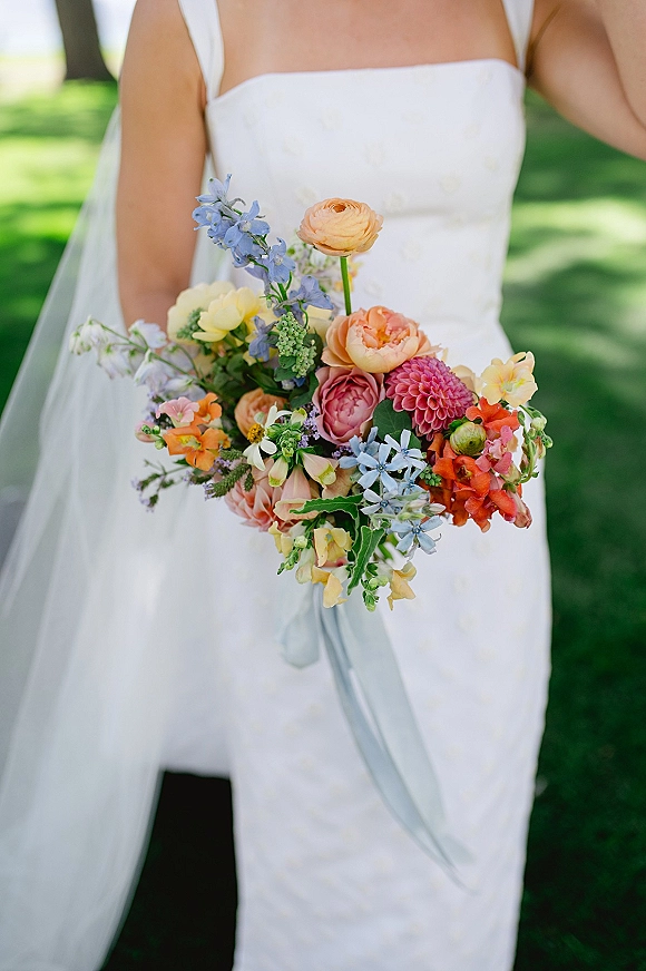 Bridal bouquet, colorful wedding bouquet with ranunculus, dahlia and delphinium, tied with pastel ribbon, held over a white dress on green lawn