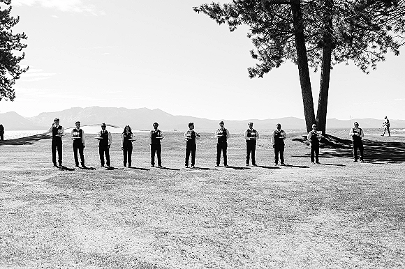 Wedding waiters offering champagne service wedding drinks, standing in vests with tray of flutes on a lakeside lawn with mountains and pines behind