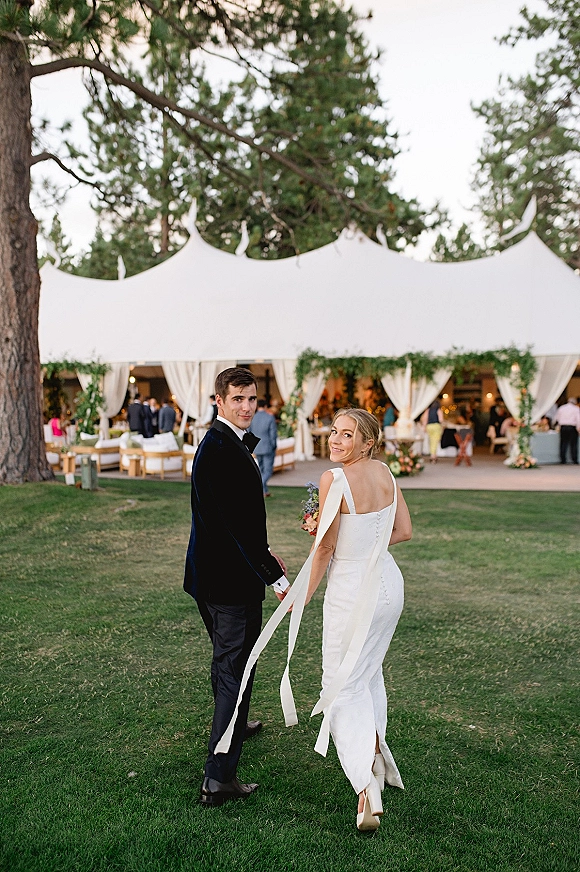 Newlywed couple portrait with bride and groom walking away hand in hand, bride looking back, bouquet ribbons flowing at a white tent reception on the lawn