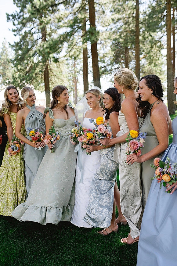 Bridesmaid group photo with bride and bridesmaids in mismatched satin dresses holding colorful bouquets on a lawn before pine trees in daylight