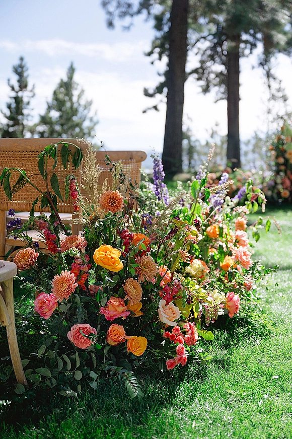 Ceremony floral decor of grounded ceremony florals with roses and dahlias, lush greenery around a rattan chair on a pine-lined lawn