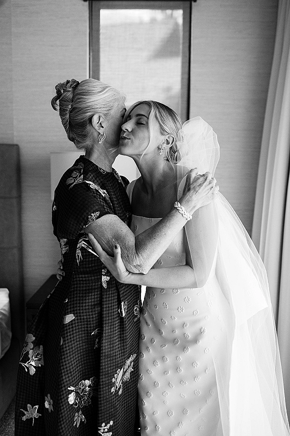 Getting ready moment as mother and bride hug, bride in veil and sleeveless wedding dress by window light in a hotel room