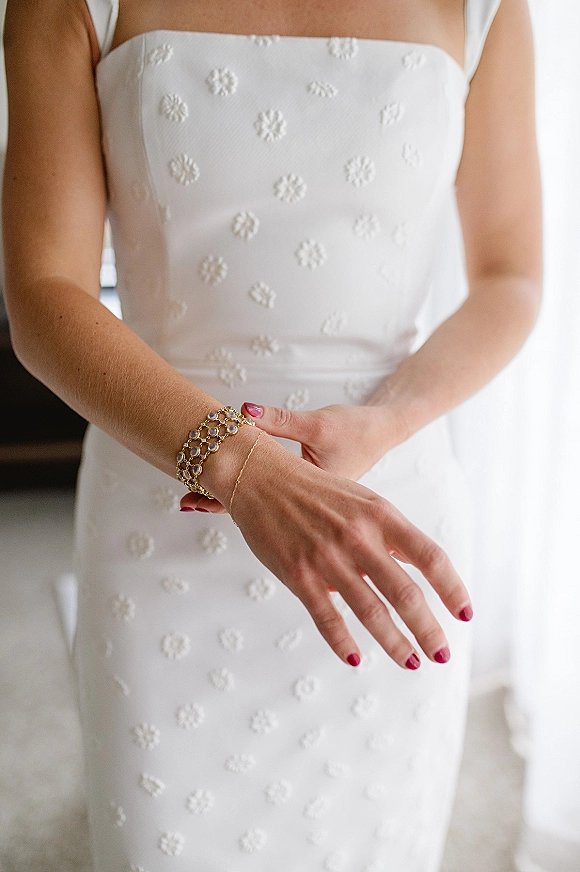 Wedding dress close-up of a strapless wedding dress with floral appliqué bodice, bride’s manicured hands and gold bracelet in window light