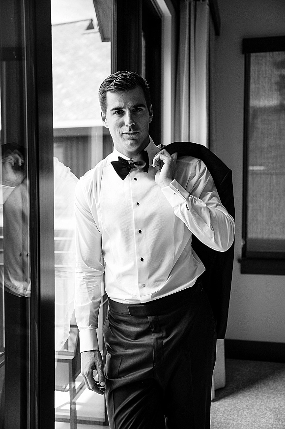 Groom portrait in a tuxedo jacket and black bow tie, standing by a window near a glass door with framed artwork and balcony railing
