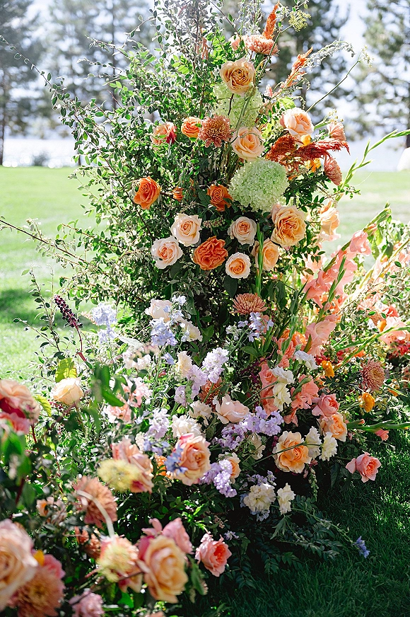 Wedding floral arrangement with peach roses and hydrangea blooms, lavender flowers, and greenery on a lakeside lawn under daylight sky