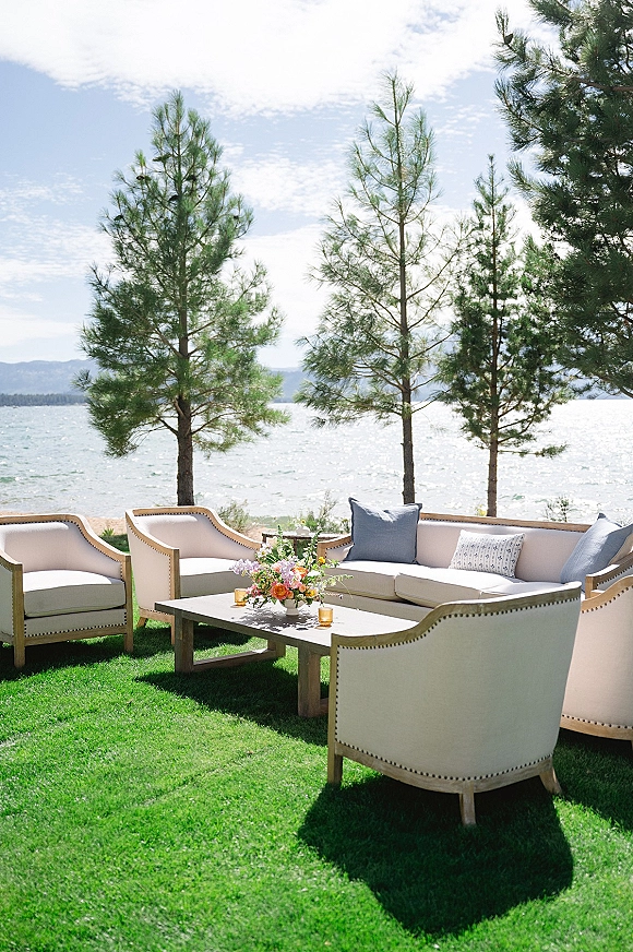 Outdoor lounge seating with upholstered sofas and chairs, throw pillows, and a floral centerpiece on a coffee table beside a lake
