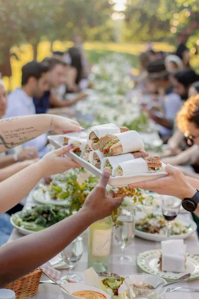 Wedding reception dinner at an outdoor wedding dinner with guests passing a sandwich platter along a long banquet table under trees