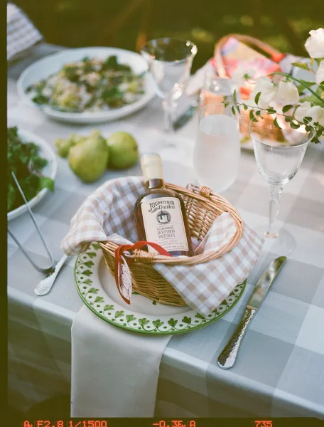 Reception tablescape with gingham wedding table linens, wicker basket centerpiece, pear and florals, and mini liquor bottle favor on lawn at dusk