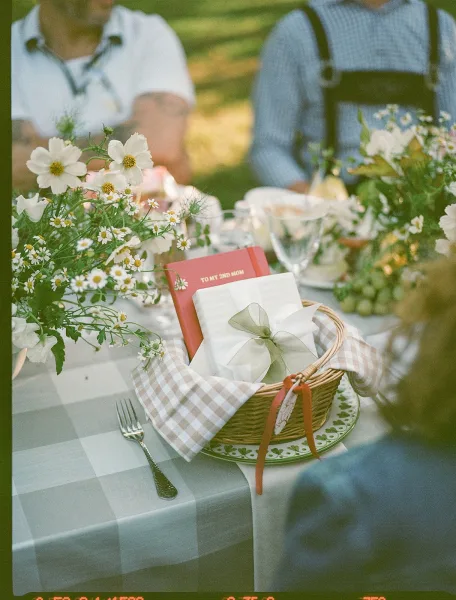 Reception tablescape with an outdoor reception table, blue gingham cloth, daisy wildflowers, woven favor basket, and guest book on lawn
