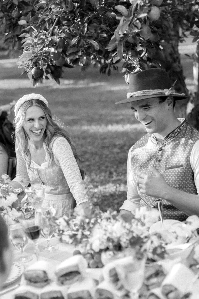 Wedding reception moment of bride and groom laughing at sweetheart table with floral centerpieces under fruit tree branches on lawn