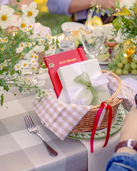 Wedding place setting with wicker basket holding a napkin tied with green ribbon, set on checked tablecloth with fruit and flowers outdoors