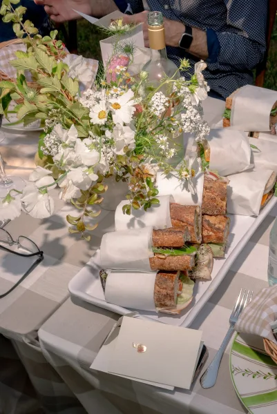 Wedding catering table with a wedding sandwich platter, parchment-wrapped baguette sandwiches and wildflower centerpiece on grass with seated guests nearby