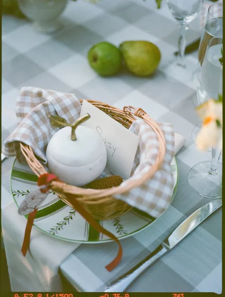 Wedding place setting with gingham wedding tablescape details: basket place card, pear on layered plates, silverware and glasses on white linen