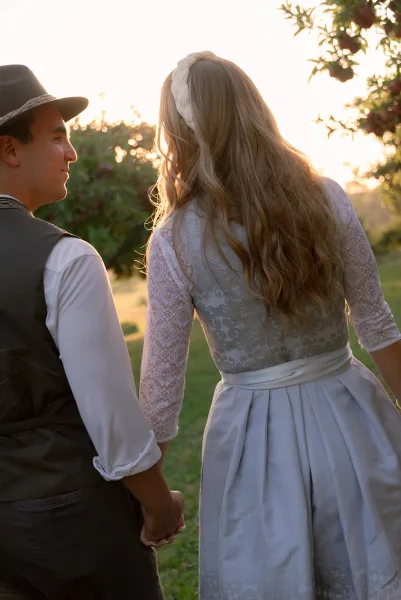 Couple portrait of bride and groom holding hands, walking away in a grassy orchard at sunset, bride in lace wedding dress with sash