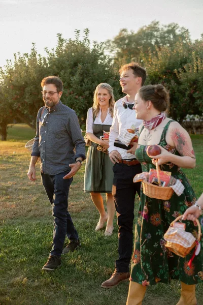 Wedding guests carrying wicker baskets with gingham napkins and bottled drinks as they walk through a grassy field in golden hour light