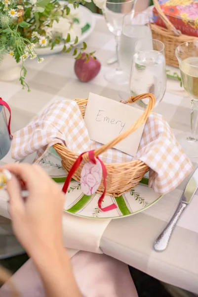 Reception tablescape with a wedding place card beside a woven favor basket, gingham napkin, patterned plates, glassware, and greenery centerpiece