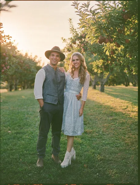 Couple portrait in an apple orchard at sunset, bride in short lace-sleeve dress with blue sash and ankle boots, groom in vest and hat