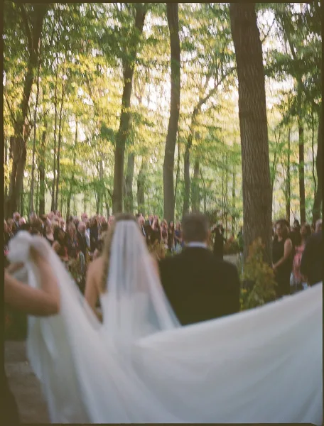Processional as bride walking down aisle in flowing wedding gown with bridal veil, guests watching along a sunlit forest aisle runner