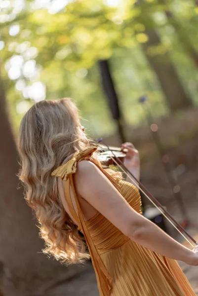 Wedding musician, wedding violinist playing violin with bow in a pleated tie-strap dress, long wavy hair backlit by sunlit trees