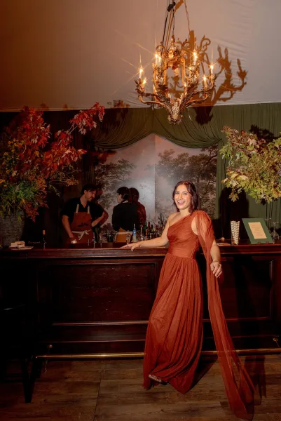 Wedding guest portrait in a rust bridesmaid dress with chiffon drape, posing by an indoor bar under a chandelier with cocktail glassware