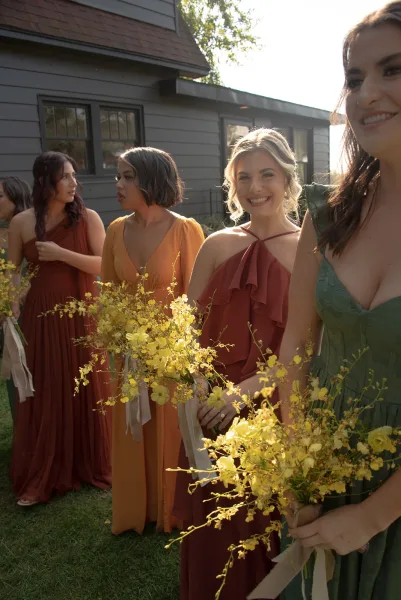 Bridesmaid portraits of bridesmaids holding bouquets, lined up laughing in mix-and-match dresses with yellow bouquets and ribbon streamers outside a sunlit house