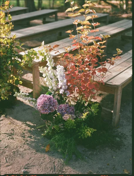 Ceremony aisle florals with hydrangeas, tall white blooms, and greenery accented by fall foliage beside wooden benches on sandy garden ground