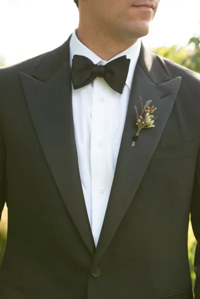 Groom portrait in a black tuxedo jacket with bow tie and boutonniere, standing in sunlight against soft outdoor greenery background