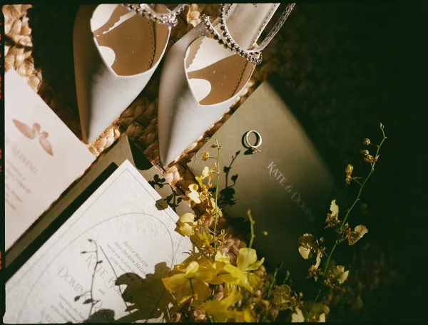 Wedding details flat lay with wedding invitation flat lay, white bridal heels, rings, vow book, envelope, and yellow flowers on a dark surface