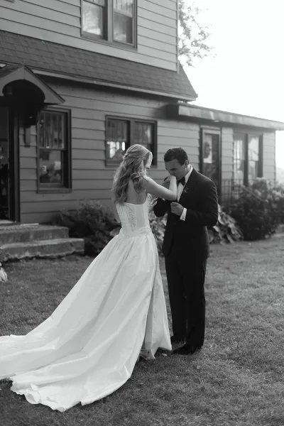 First look moment as groom kissing bride’s hand while holding hands, her strapless gown and long train on a lawn by house steps