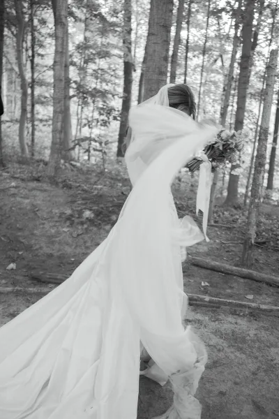 Bridal portrait of a bride walking away with bouquet and veil, her long dress train trailing along a forest dirt path among trees