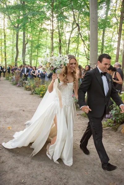 Wedding recessional as bride and groom walk the aisle, bride lifting bouquet overhead, guests cheering along a forest dirt path