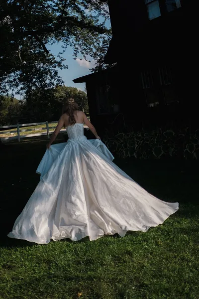 Bridal portrait of a bride in a strapless ball gown with a long train, walking across a grassy lawn by a rustic barn and fence
