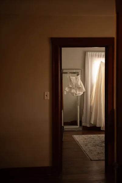 Wedding dress detail with lace bodice hanging by a window, reflected in a floor mirror in a softly lit bedroom getting-ready space