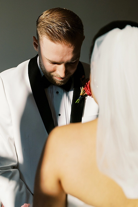 Ceremony moment as groom looks down in an emotional moment, wearing a white tuxedo jacket with boutonniere beside bride’s veil against a neutral wall