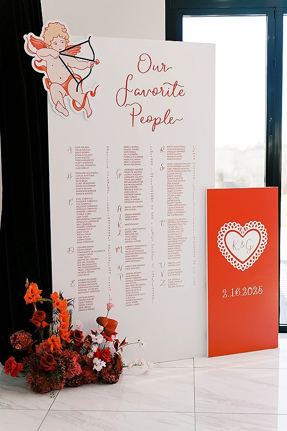Wedding seating chart with alphabetical list in script typography, paired with a red welcome sign and roses by a sunlit window