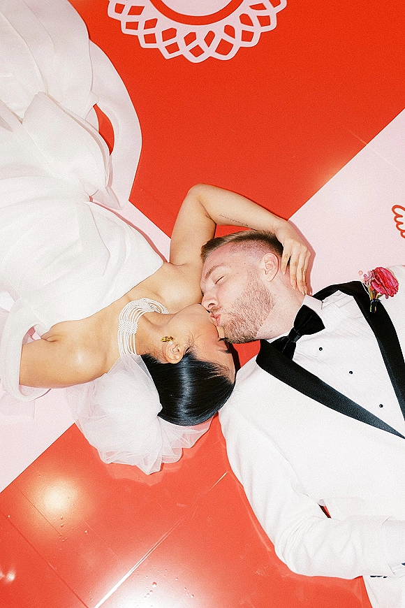 Wedding kiss portrait in an overhead wedding photo of bride and groom lying on a red floor with white graphic pattern, veil and tuxedo details