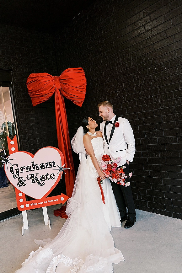 Couple portrait of bride and groom posing as she looks up at him, holding a red-pink bouquet before a black brick wall backdrop