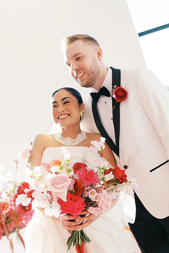 Couple portrait of bride and groom, bride holding a red and pink bouquet in veil and white dress, groom in white tuxedo by window light