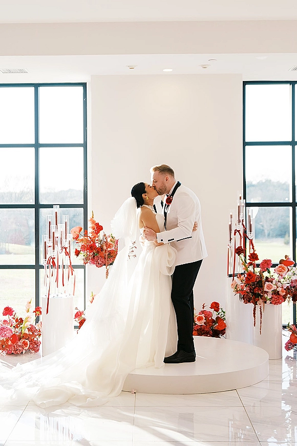 Wedding kiss as bride in strapless gown and long veil leans into groom in white tux on a circular stage amid florals and candles