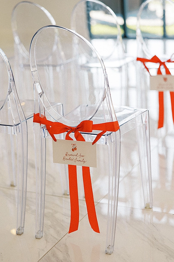 Ceremony reserved seating with reserved seat wedding sign cards on clear acrylic chairs, tied with red ribbon bows in an indoor venue