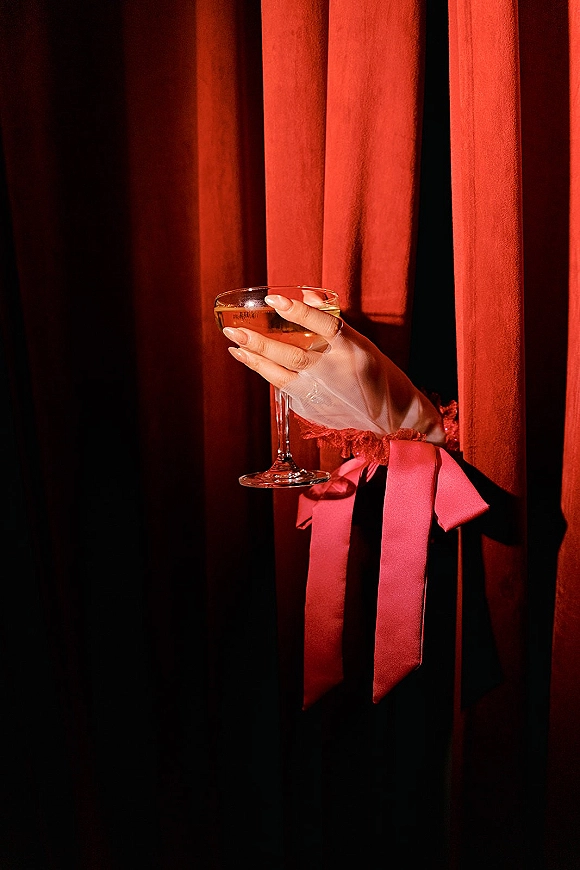 Champagne coupe held in sheer gloves with a red ribbon bow and fresh manicure, set against moody red velvet curtains and shadows