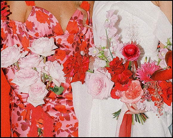 Wedding bouquet of pink and red roses, ranunculus, and sweet peas with greenery held against a dark backdrop, gold ring visible