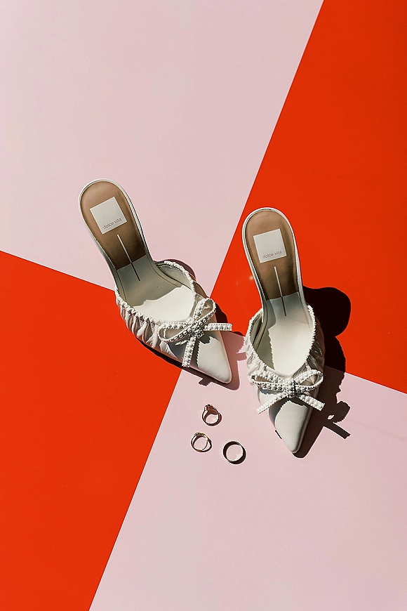 Bridal shoes flatlay of white pointed-toe mules with pearl bow details and wedding rings on a bold red and blush pink backdrop