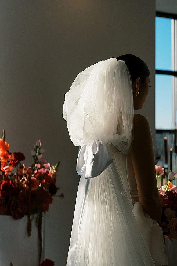 Bridal portrait of a back view bride in a strapless wedding dress with a bow wedding veil, pearl earrings, and window light indoors