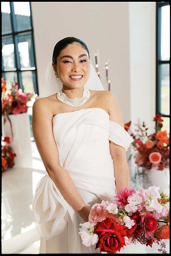 Bridal portrait of a smiling bride in a strapless wedding dress with veil, pearl necklace, and rose bouquet by large windows in daylight