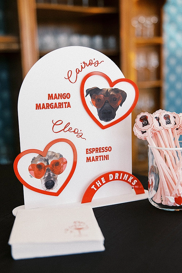 Wedding drink menu sign with red lettering and pet illustrations, heart cutouts and pink drink stirrers on bar table with glassware behind