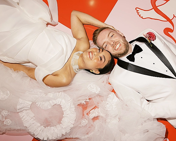 Couple portrait of bride and groom lying down, her strapless dress and tulle veil spread beside his white tuxedo on red-pink backdrop