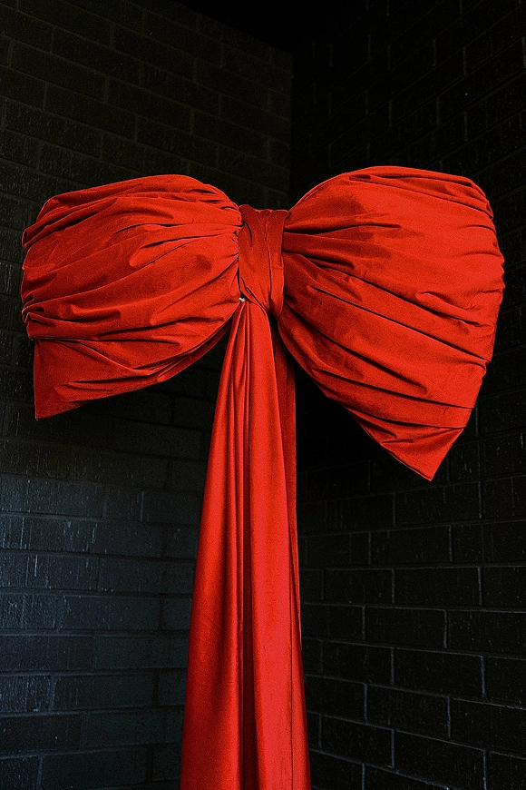 Wedding bow decor featuring an oversized wedding bow of red draped fabric hanging against a black brick wall backdrop