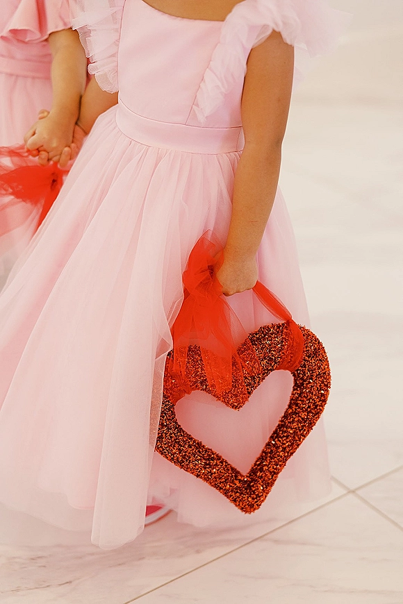 Flower girl dress with pink tulle skirt, ruffle sleeves and satin waistband, holding a red glitter heart purse with red shoes on tile floor